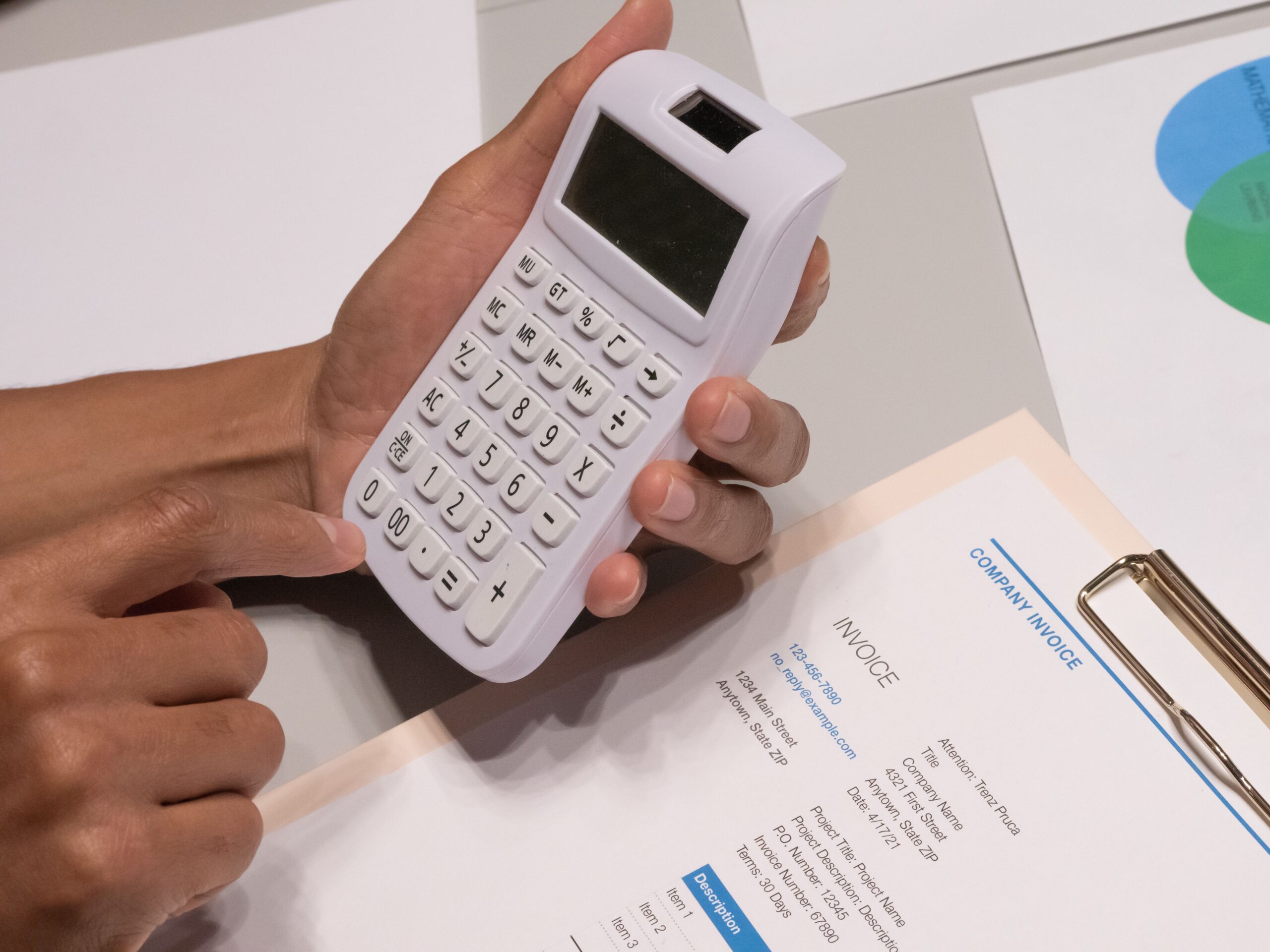 Primer plano de manos utilizando una calculadora blanca sobre una factura profesional con el texto INVOICE, representando el cálculo de importes en la facturación.