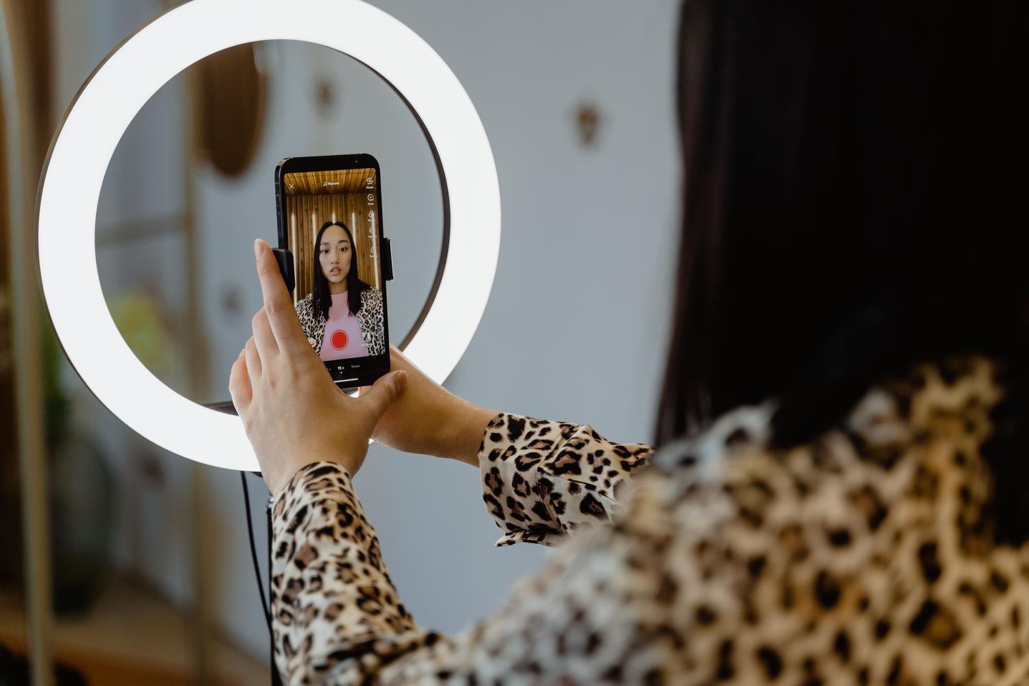 Creadora de contenido utilizando un aro de luz y su teléfono móvil para grabar un vídeo, ilustrando la estrategia de contenido UGC y Live Shopping. Creadora de contenido utilizando un aro de luz y su teléfono móvil para grabar un vídeo, ilustrando la estrategia de contenido UGC y Live Shopping.