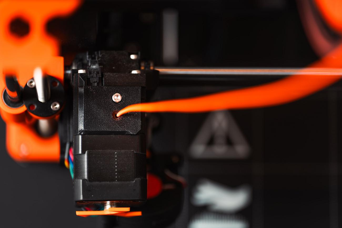 Vista detallada de una impresora 3D extrusora con filamento naranja, enfocada en el proceso de deposición de material.