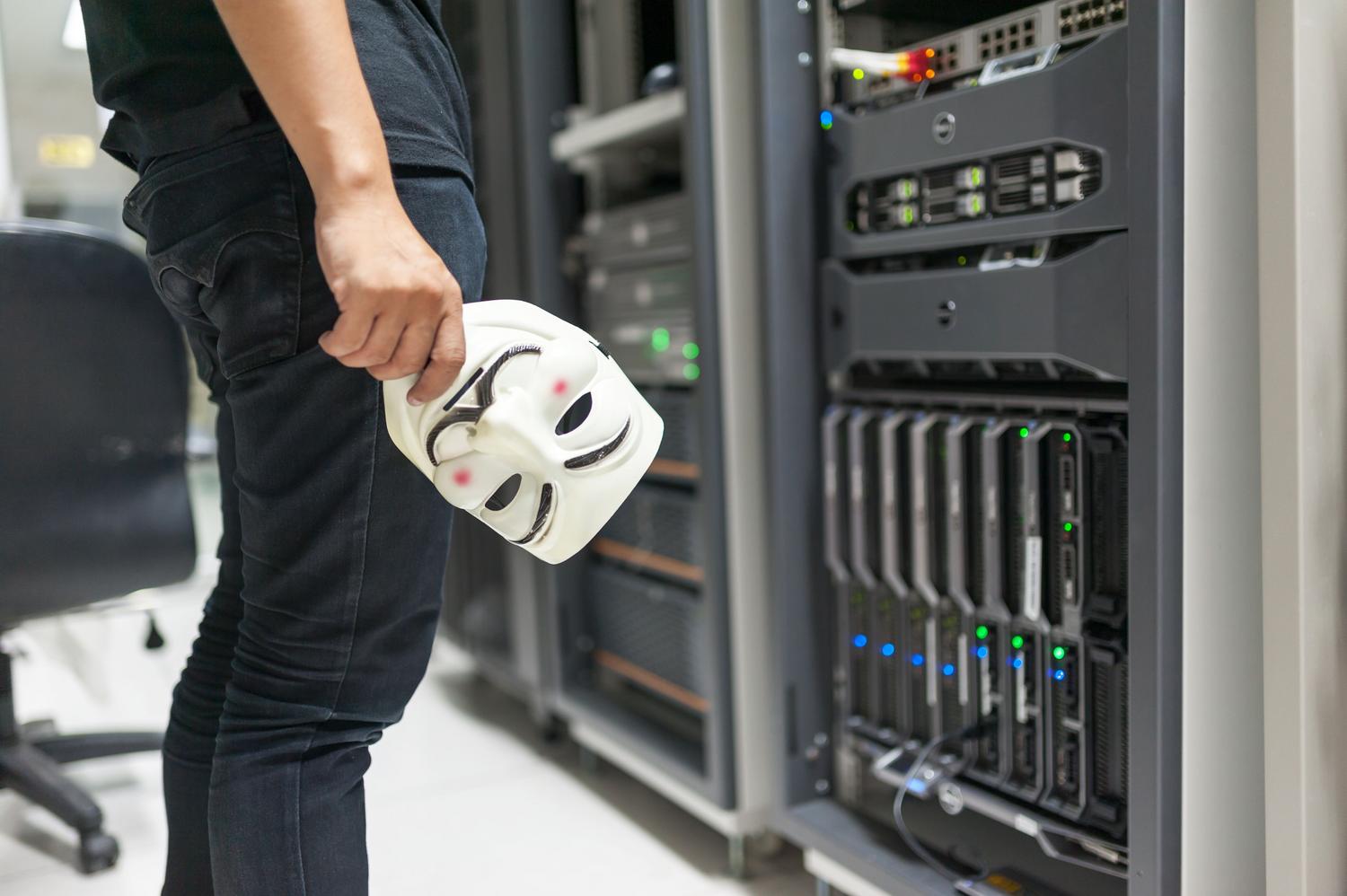 Persona con máscara de Anonymous junto a racks de servidores, simbolizando amenazas de ciberseguridad y accesos no autorizados.