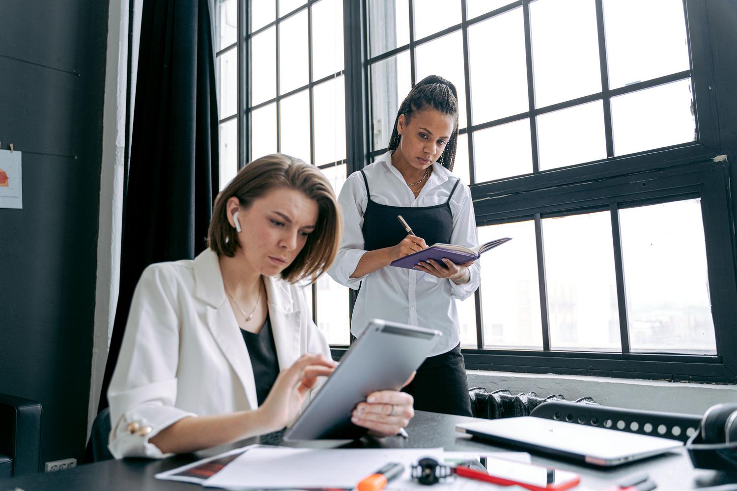 Dos mujeres profesionales colaborando en una oficina moderna con dispositivos digitales, reflejando el trabajo en equipo y la orquestación de tareas.