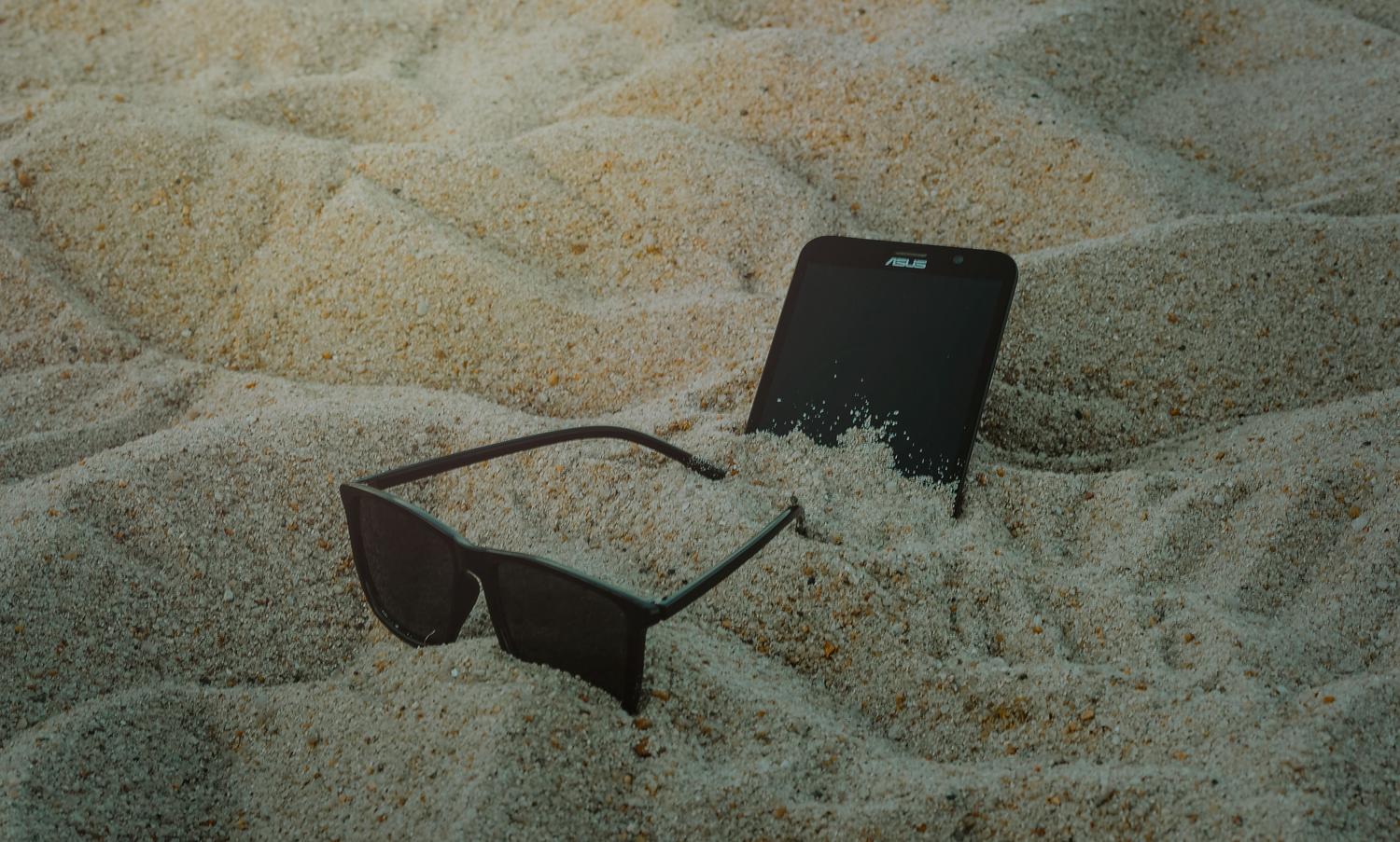 Un smartphone y gafas de sol sobre la arena de una playa, representando la exposición al calor solar.