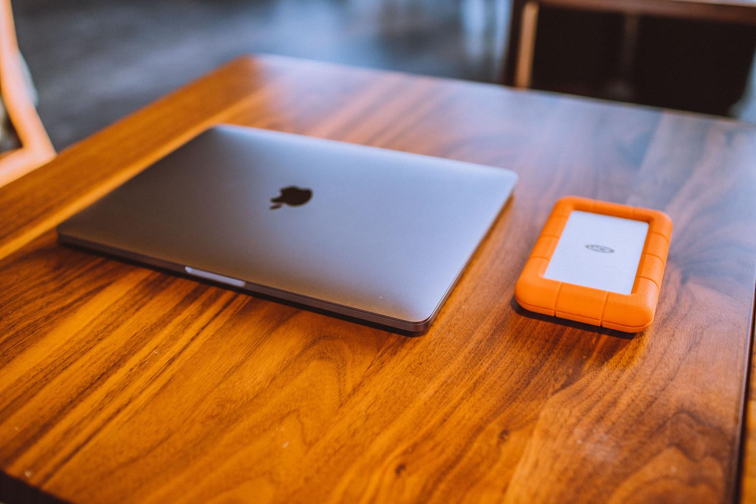 MacBook cerrado y disco duro externo Rugged sobre una mesa de madera, representando la combinación de hardware y almacenamiento externo en macOS.