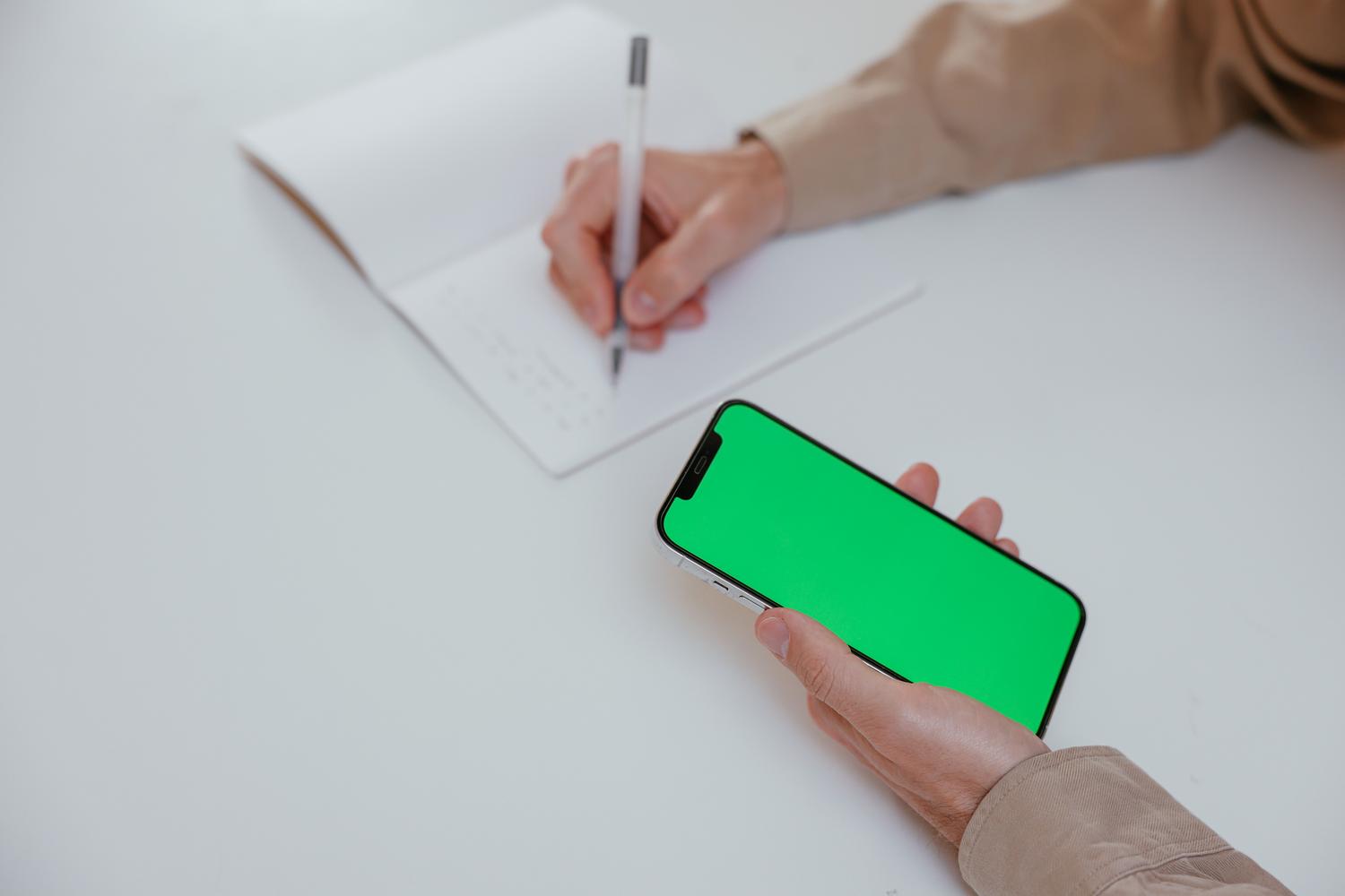 Vista cenital de una persona tomando notas en un cuaderno mientras consulta un smartphone, ilustrando la acción de escribir mientras se mira un vídeo. Vista cenital de una persona tomando notas en un cuaderno mientras consulta un smartphone, ilustrando la acción de escribir mientras se mira un vídeo.