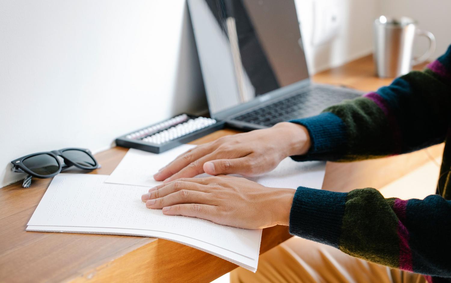 Detalle de manos leyendo Braille junto a un MacBook, destacando la compatibilidad de macOS con dispositivos táctiles.