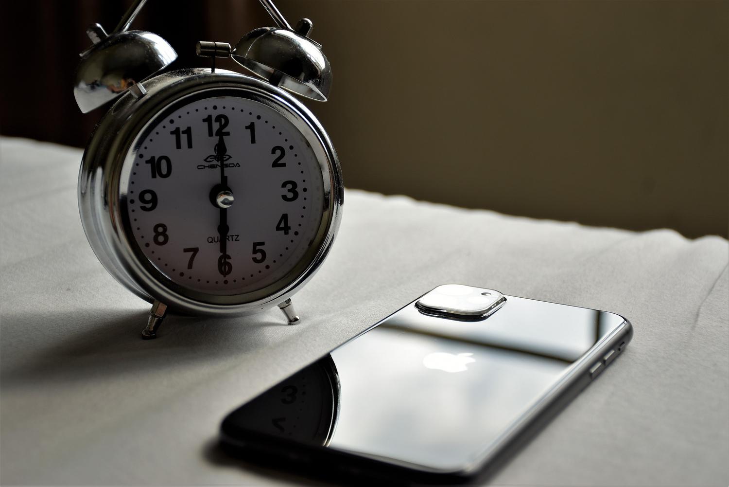 Reloj despertador clásico junto a un smartphone boca abajo sobre sábanas blancas, simbolizando que las alarmas siguen activas aunque el móvil esté en modo No Molestar.