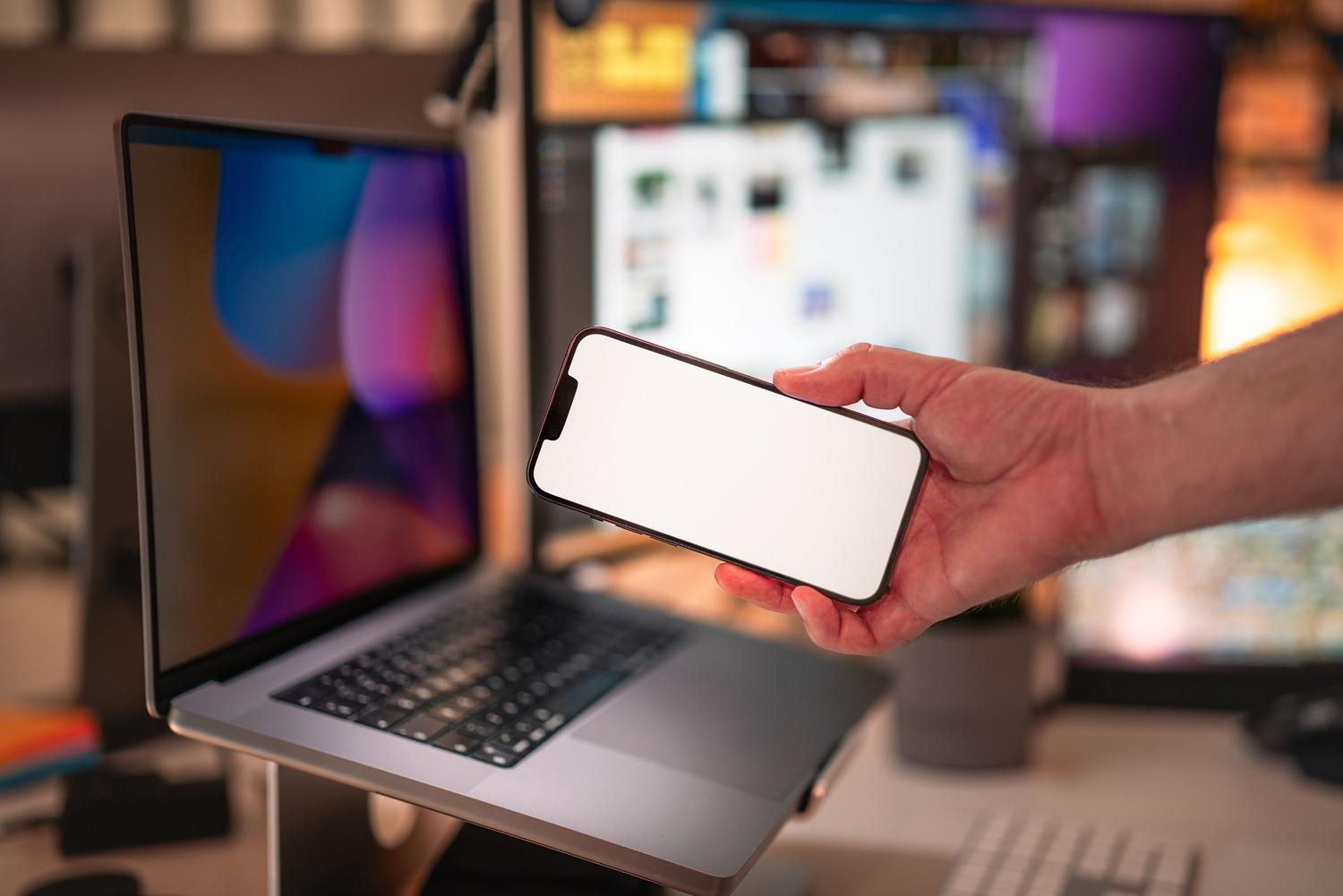 Persona utilizando un smartphone frente a un portátil en un espacio de trabajo moderno para comparar y calibrar colores.
