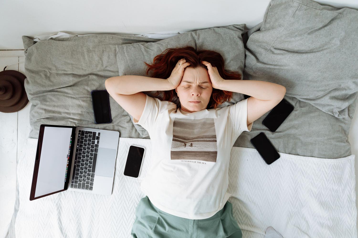 Mujer estresada en la cama rodeada de múltiples dispositivos digitales, representando la fatiga por notificaciones.
