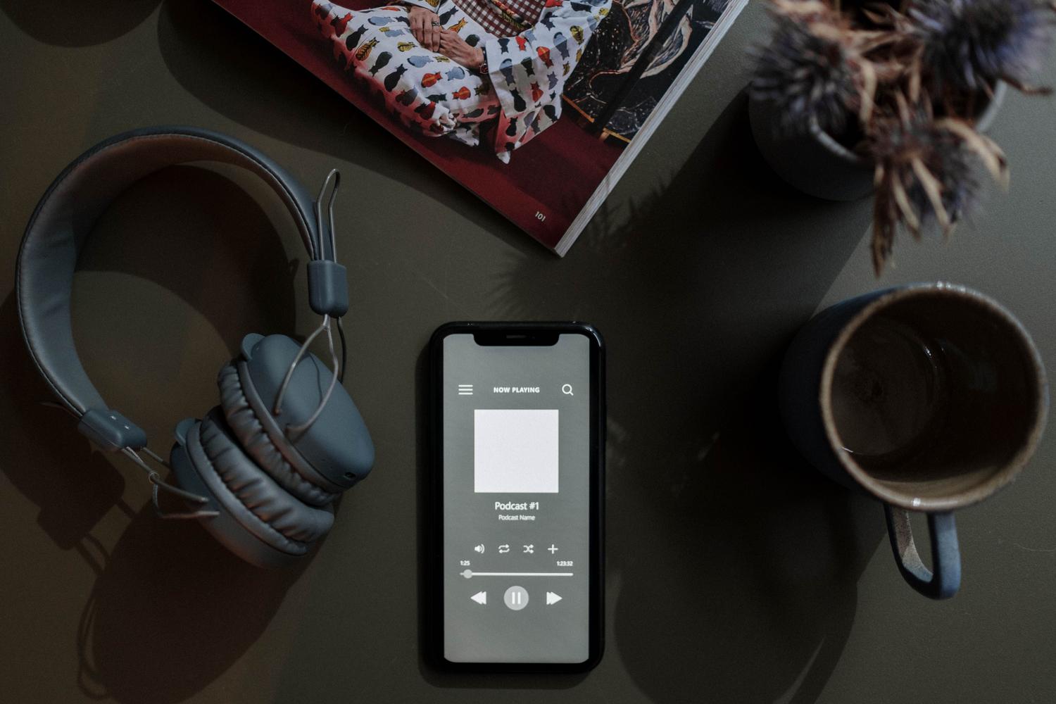 Composición cenital de un smartphone reproduciendo un pódcast junto a unos auriculares y una taza de café, ilustrando la integración de la publicidad en la rutina diaria.