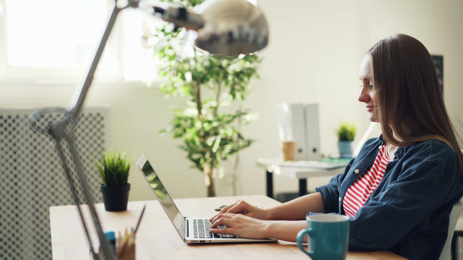 Mujer sonriendo mientras utiliza un portátil en una oficina moderna, ilustrando la experiencia de usuario al interactuar con las funciones de IA de Windows 11.