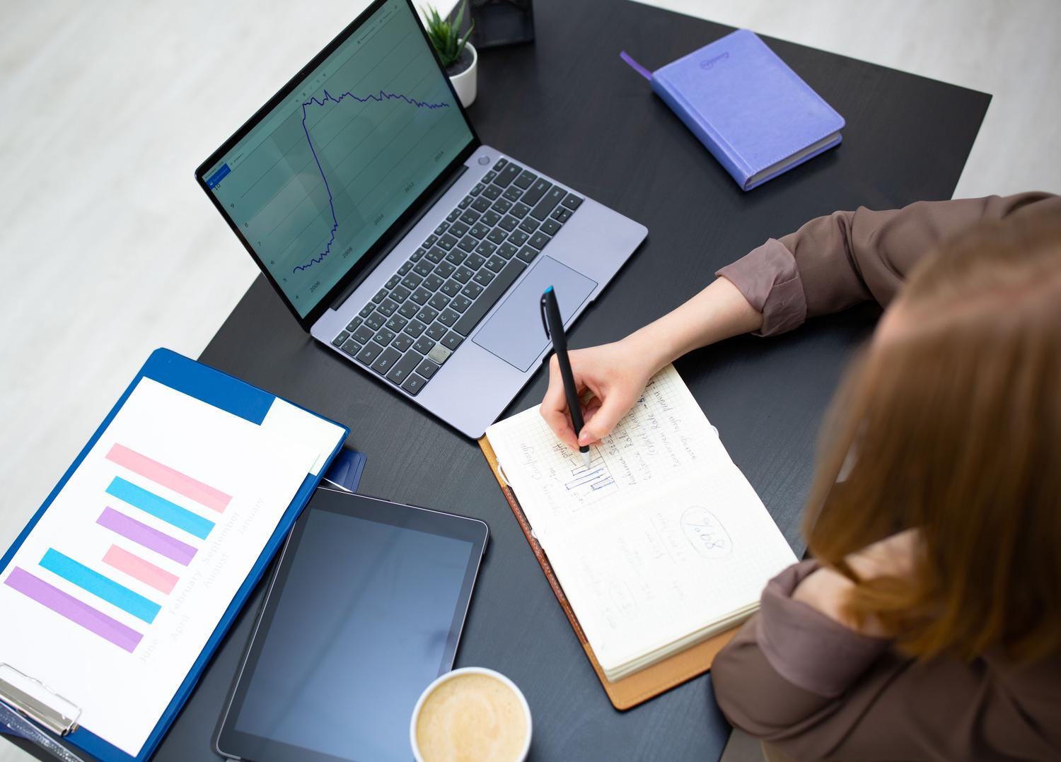 Persona organizando datos y reportes utilizando un ordenador portátil y notas manuales.