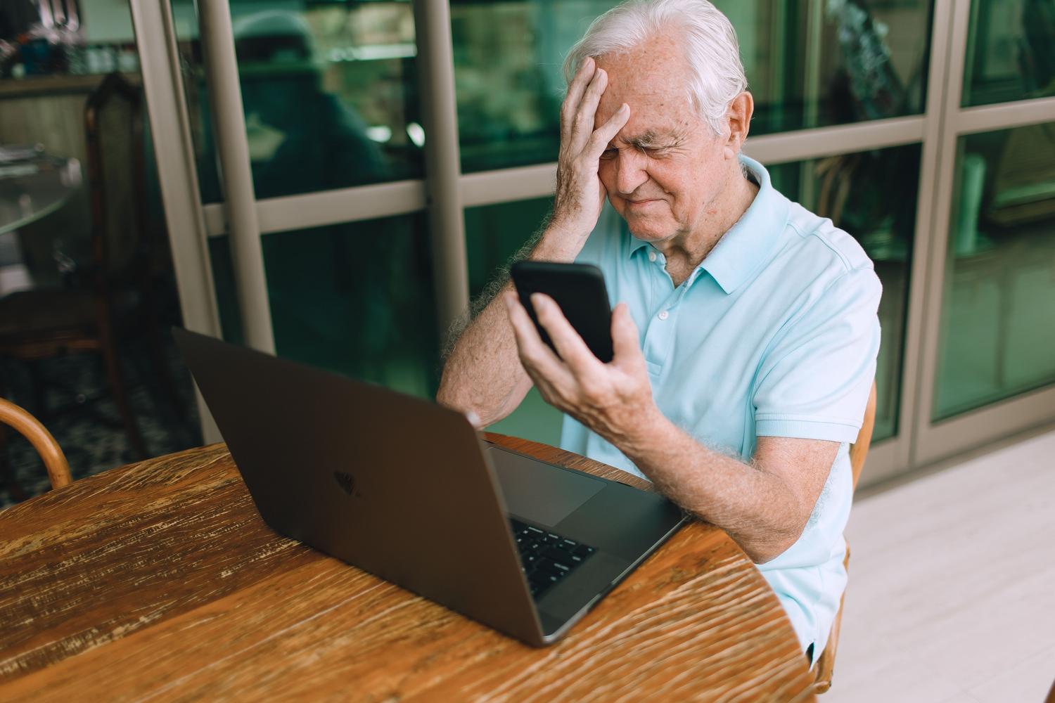 Hombre frustrado intentando solucionar problemas técnicos con su teléfono y ordenador portátil en casa