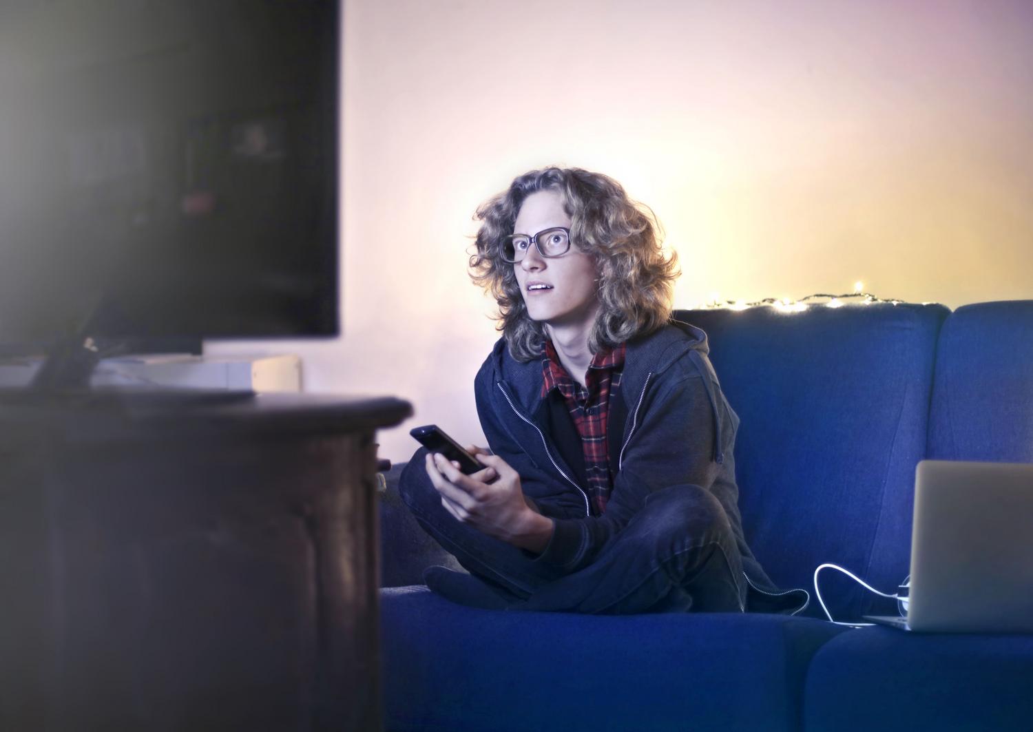 Persona sentada en un sofá de una sala de estar utilizando un smartphone mientras mira una televisión, ilustrando la conexión de dispositivos móviles mediante AirPlay.