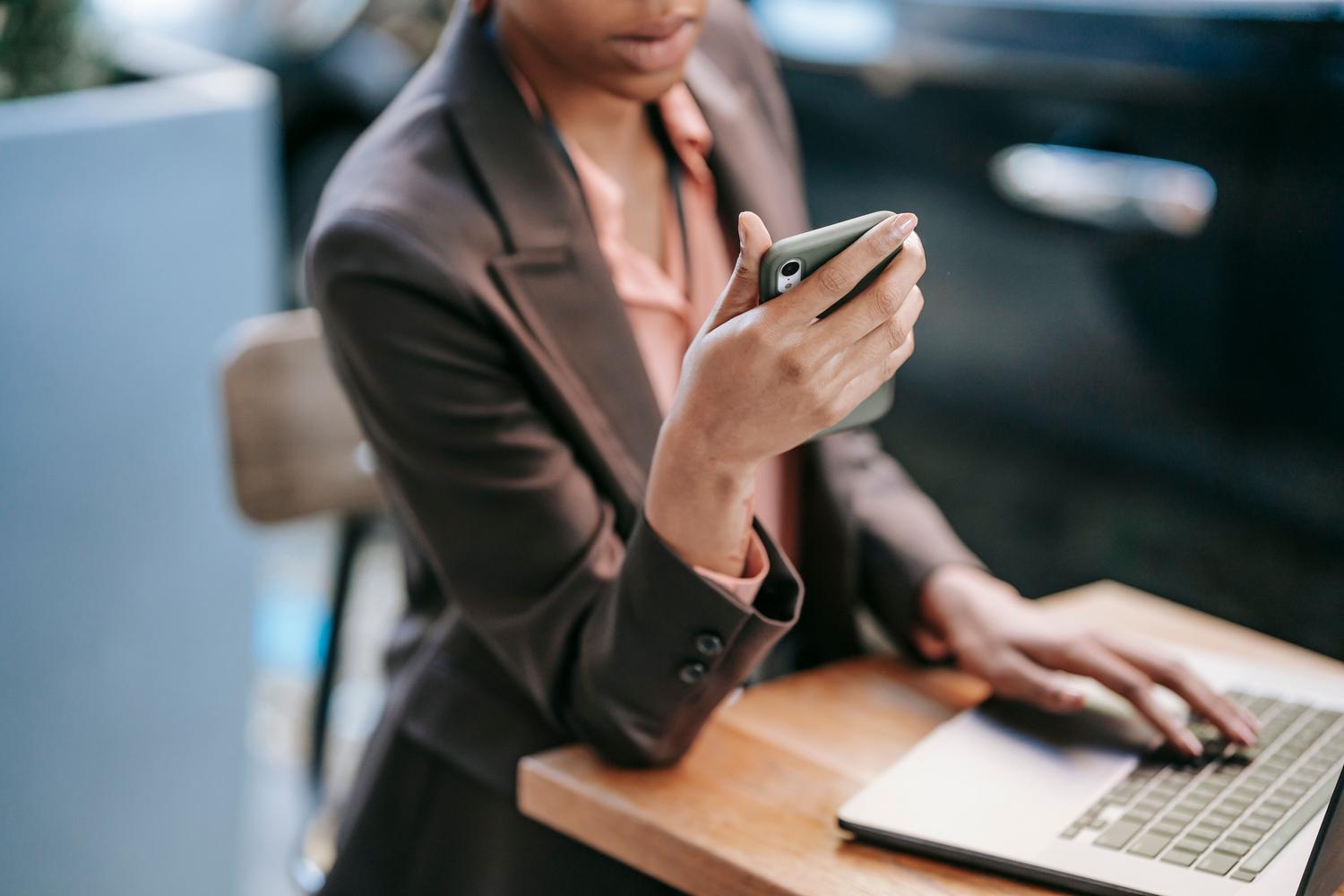 Mujer con vestimenta profesional utilizando un smartphone y un ordenador portátil al mismo tiempo, mostrando una gestión eficiente de tareas digitales.