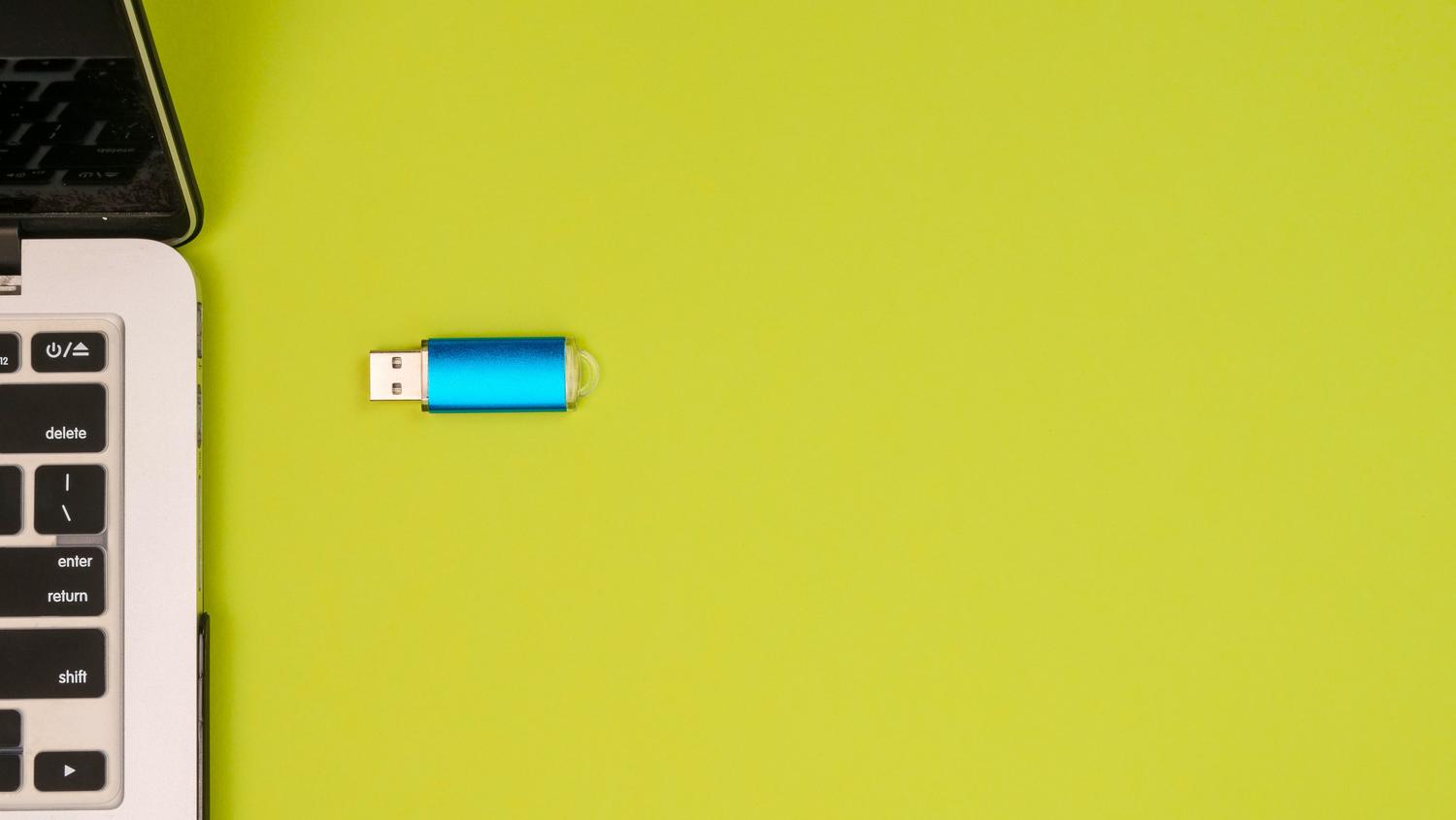 Pogled odozgo na metalik plavi USB flash pogon pored prijenosnog računala na živopisnoj žutoj pozadini, koji predstavlja modernu tehnologiju.
