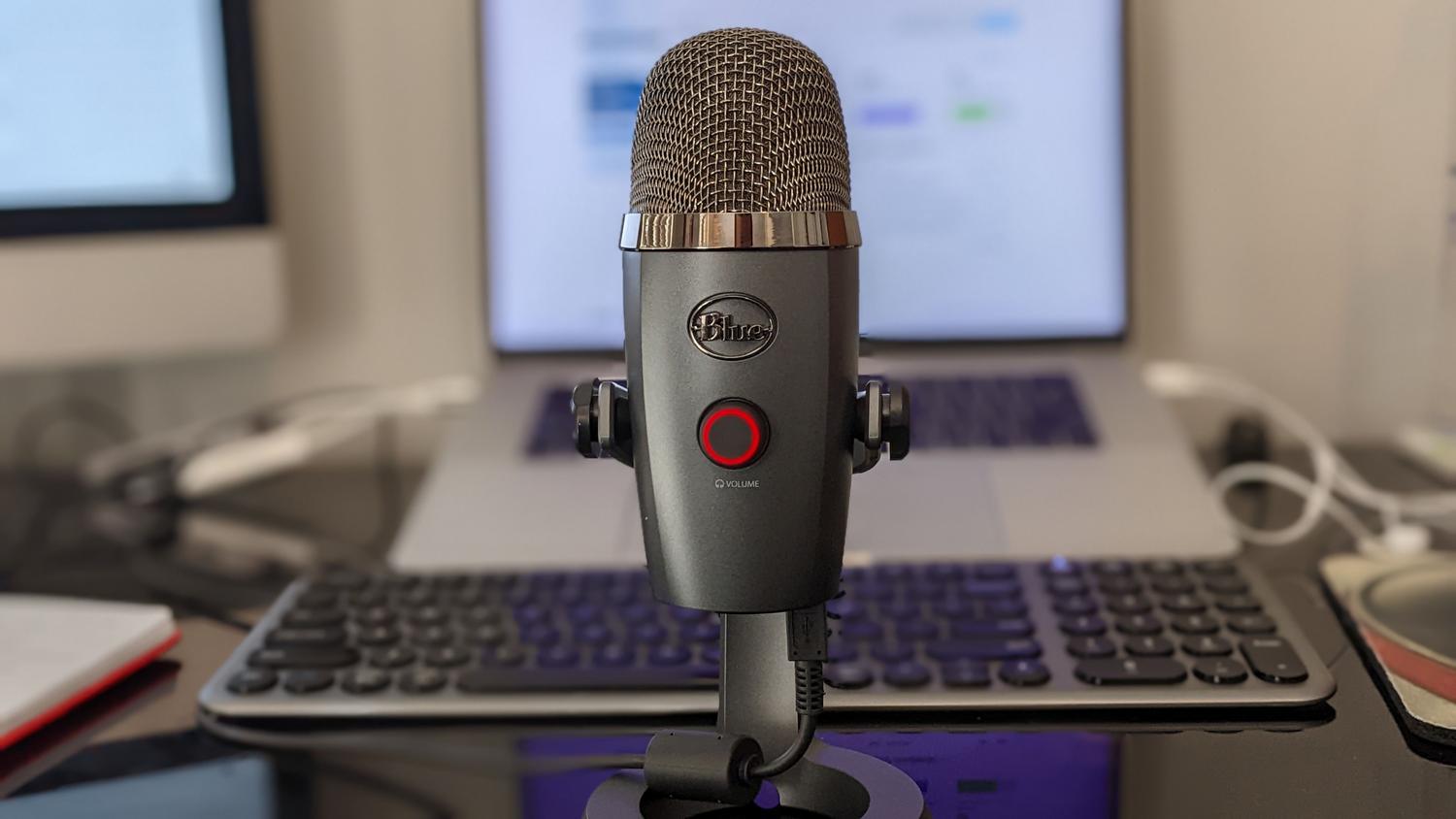 Setup de grabación de podcast casero con un micrófono Blue Yeti, una laptop y teclado en un escritorio ordenado.