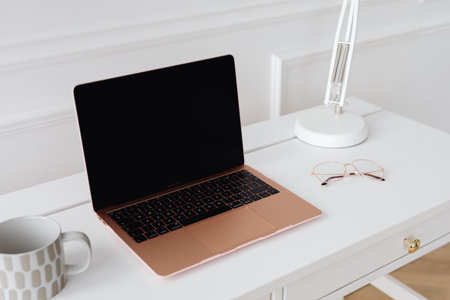 Entorno de trabajo luminoso y profesional con un MacBook, gafas y una lámpara sobre una mesa blanca.