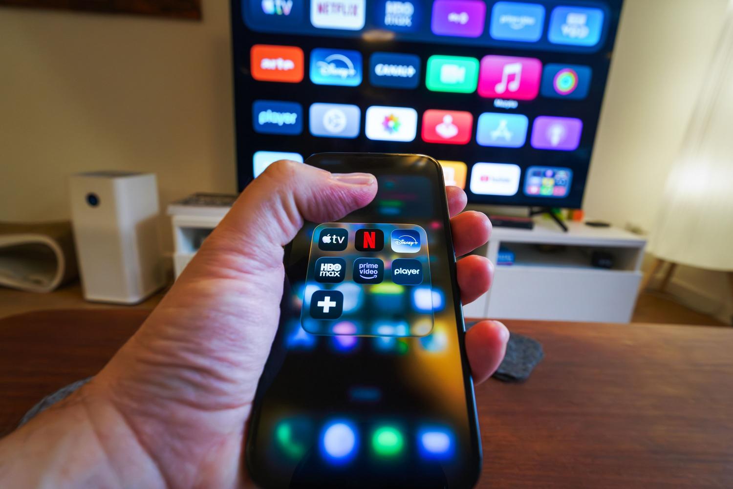 Imagem em close-up de um telefone celular exibindo aplicativos de streaming com uma televisão ao fundo, ilustrando a transmissão de conteúdo (Cast).