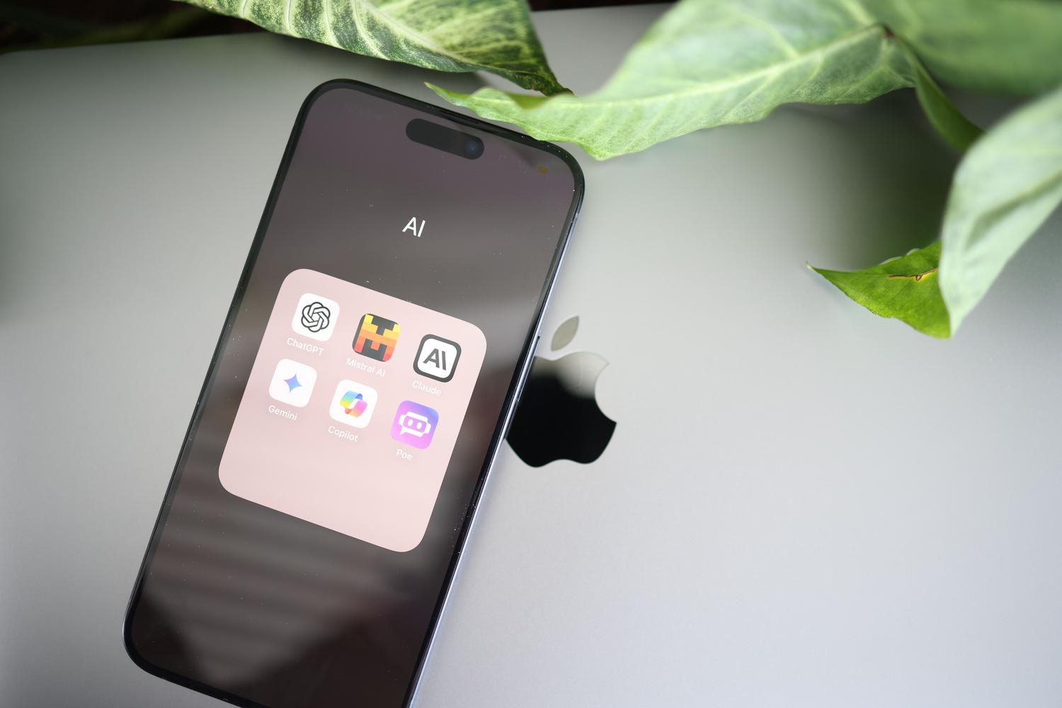 Un smartphone mostrando aplicaciones de IA apoyado sobre un ordenador portátil en un escritorio blanco con plantas verdes, representando la creación de apps con inteligencia artificial.