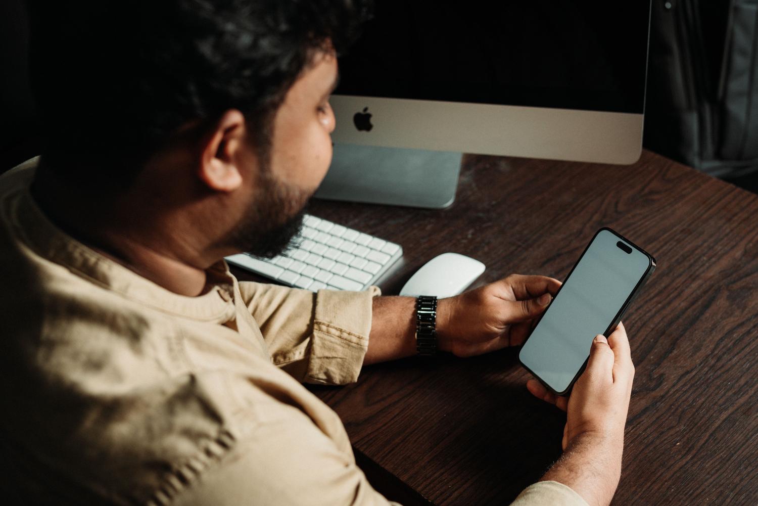 Hombre profesional trabajando en su escritorio coordinando tareas entre un ordenador de sobremesa y su smartphone.