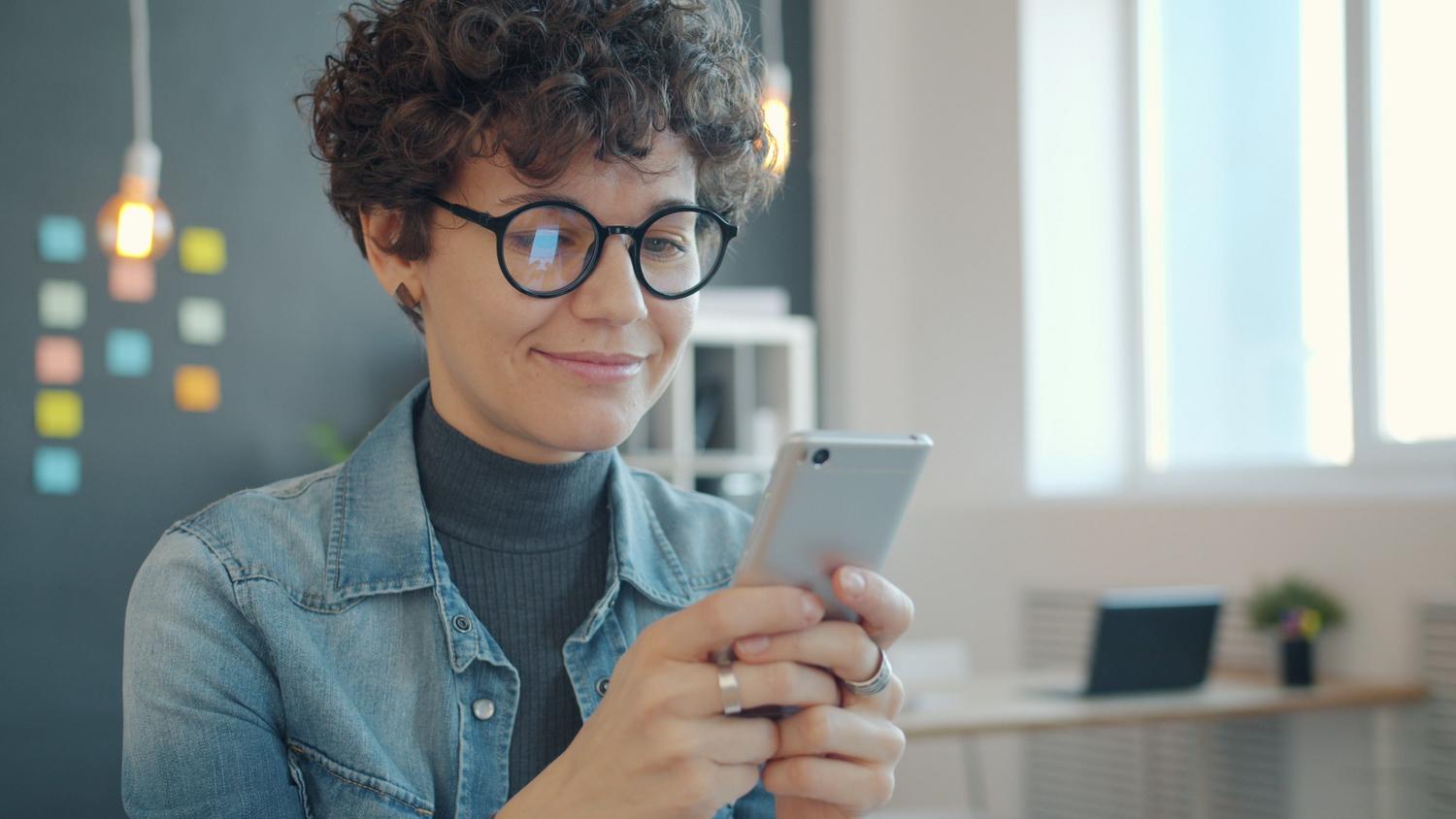 Profesional joven utilizando su smartphone en una oficina moderna y luminosa con notas adhesivas de organización al fondo.