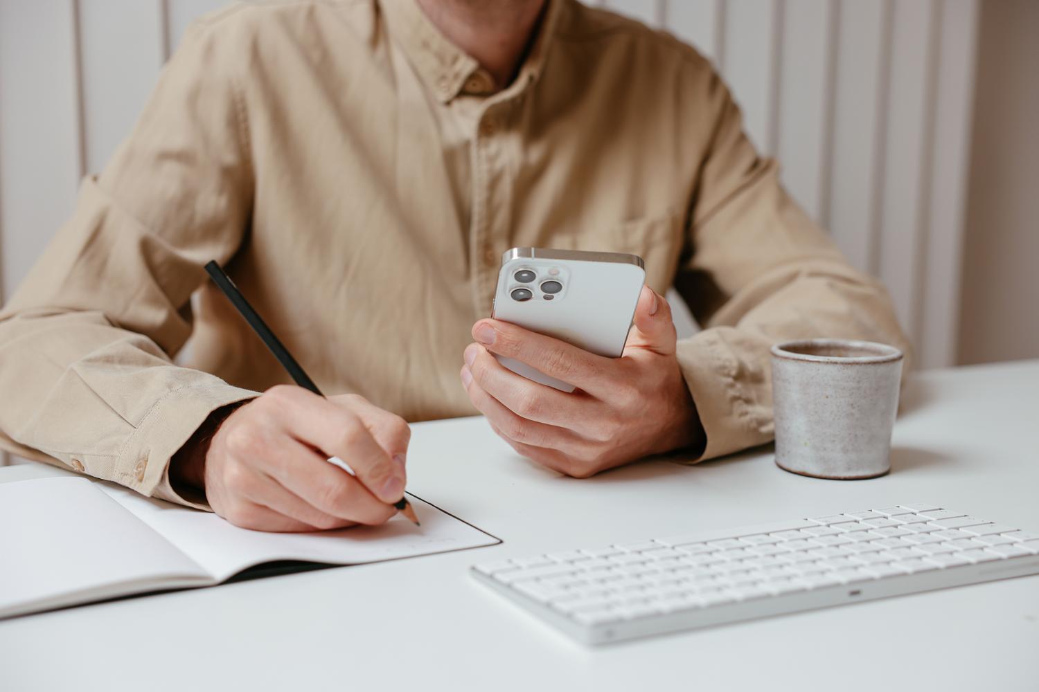 Persona realizando multitarea escribiendo en un cuaderno mientras consulta su smartphone en un escritorio blanco.
