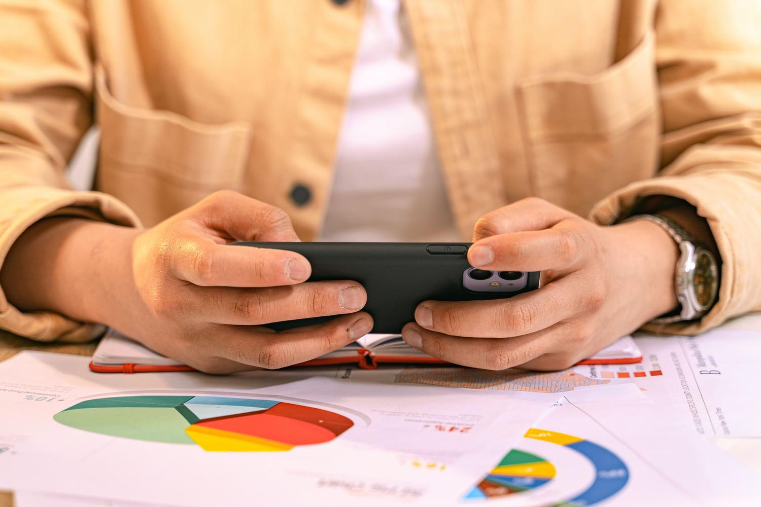 Persona analizando gráficos de datos en papel mientras gestiona tareas en su smartphone móvil.