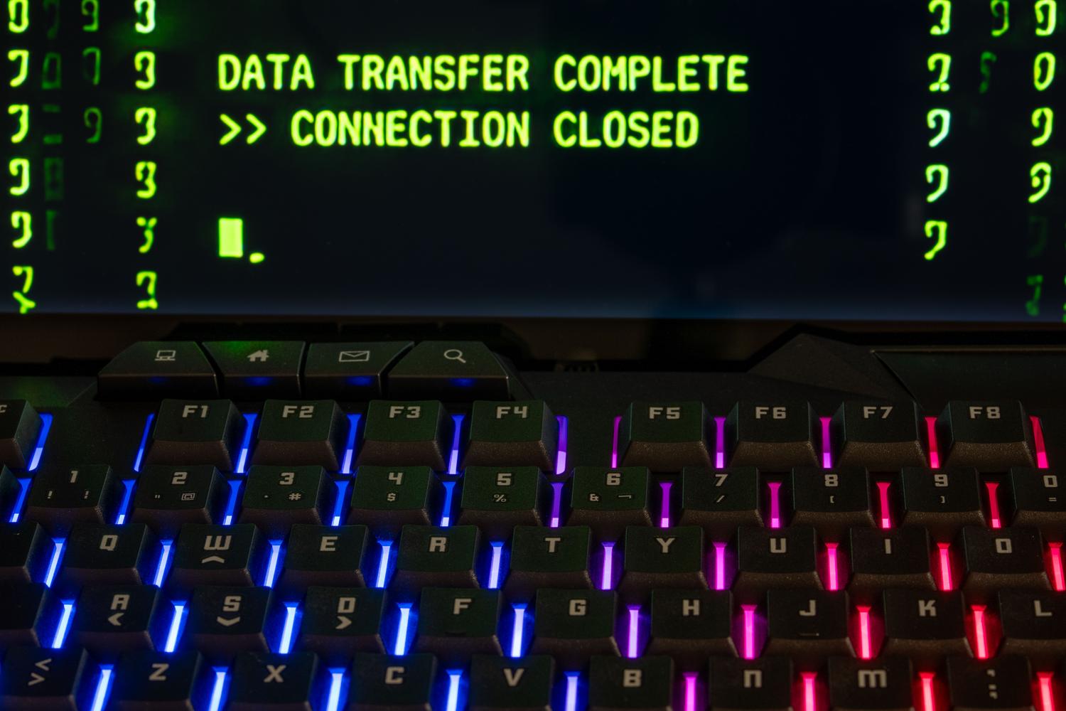 Teclado mecánico RGB y monitor con el mensaje 'DATA TRANSFER COMPLETE', ilustrando la ejecución exitosa de un script de sincronización de archivos.