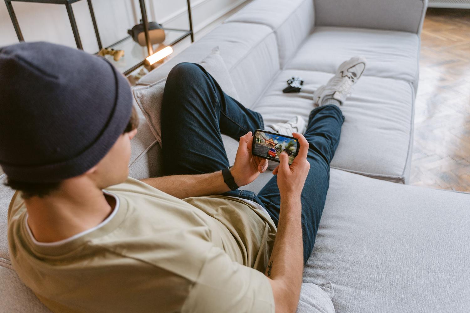 Hombre joven jugando cómodamente a un videojuego en su móvil desde el sofá. Hombre joven jugando cómodamente a un videojuego en su móvil desde el sofá.