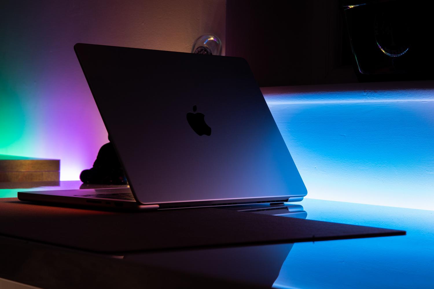 MacBook plateada abierta sobre un escritorio oscuro, iluminada por luces neon azules y magenta, creando una atmósfera de gaming moderna.