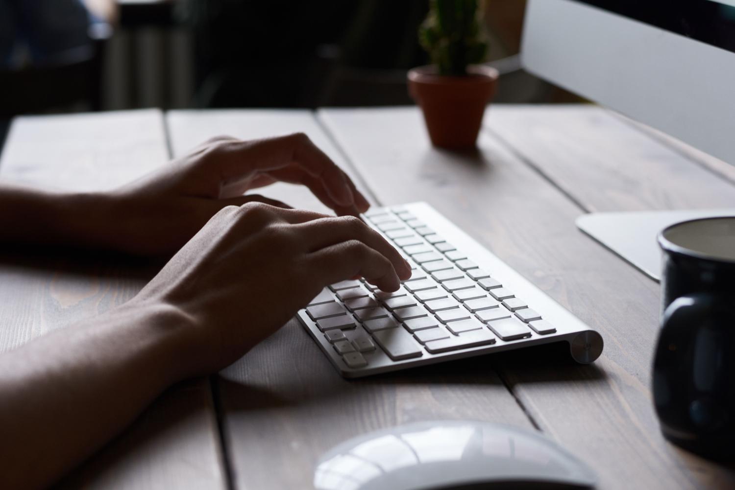 Manos escribiendo en un teclado Magic Keyboard de Apple en un escritorio de madera moderno y minimalista.