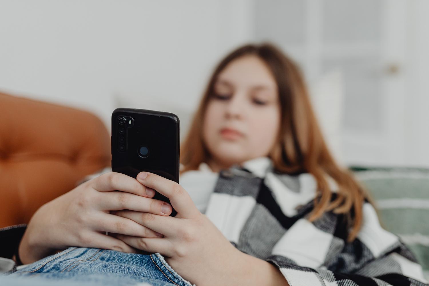 Primer plano de un teléfono móvil con una adolescente desenfocada al fondo, simbolizando la desconexión de la realidad.