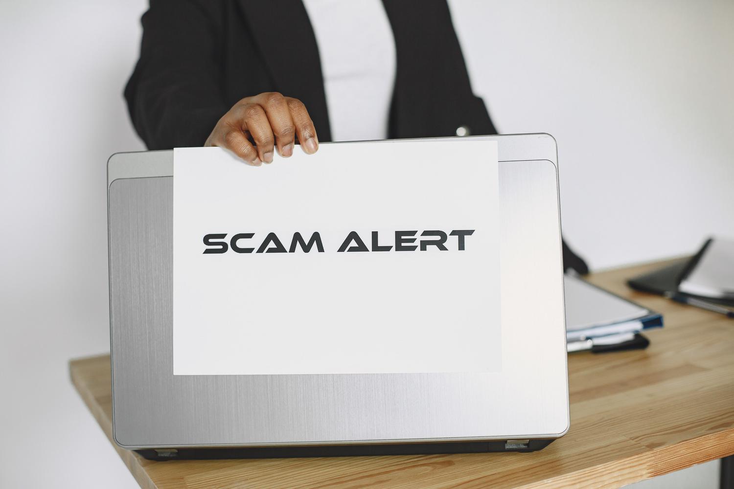 Persona en entorno profesional sosteniendo un aviso de alerta de estafa (Scam Alert) sobre un ordenador