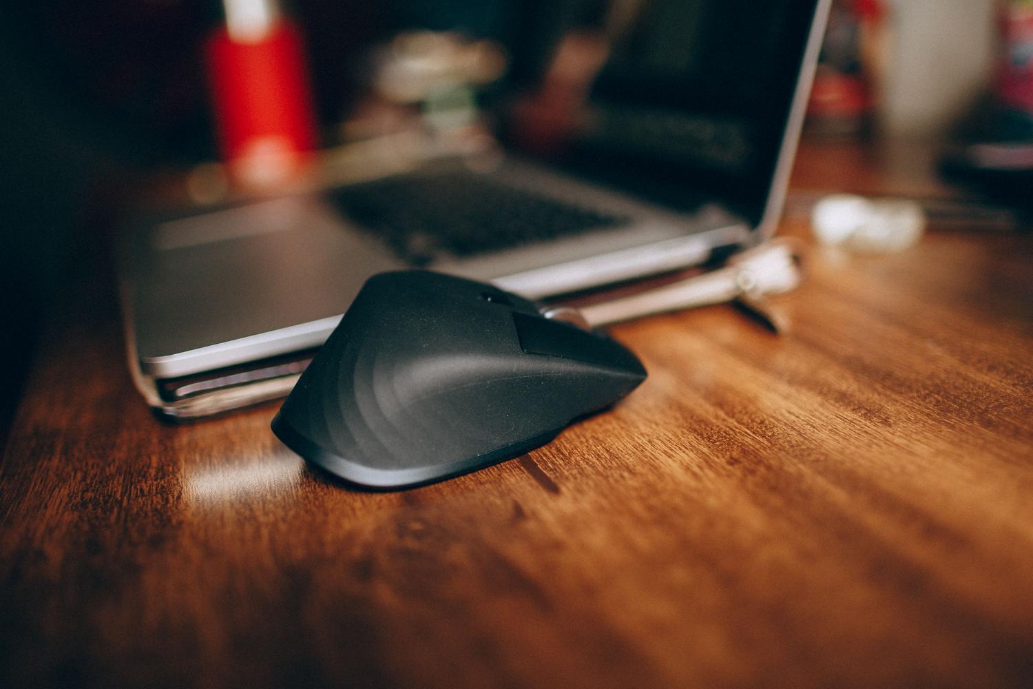 Ratón inalámbrico ergonómico negro junto a un ordenador portátil sobre un escritorio de madera.