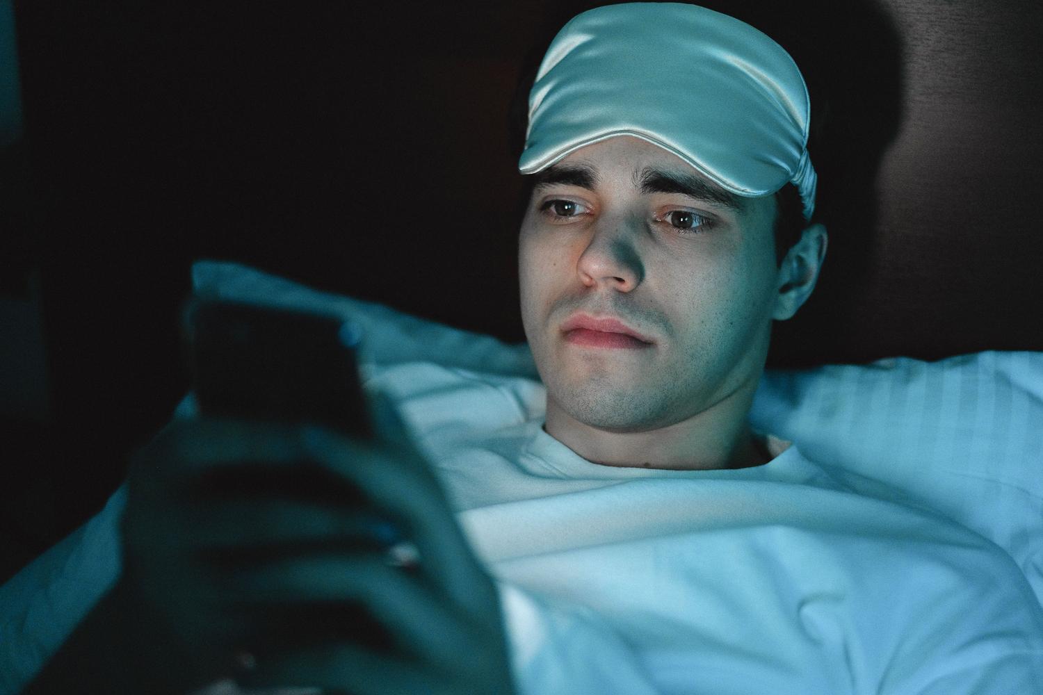 Hombre usando un antifaz para dormir mientras consulta su teléfono móvil antes de descansar.