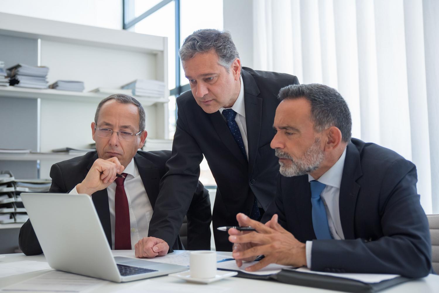 Tres ejecutivos en trajes formales colaborando en una oficina moderna, analizando datos en un ordenador portátil.
