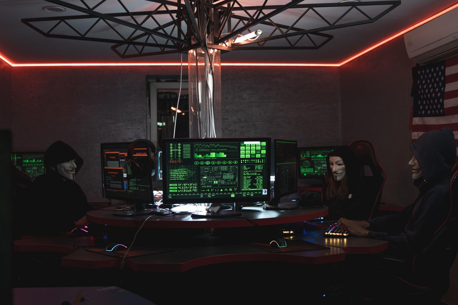 Grupo de hackers anónimos con máscaras trabajando en una sala oscura con luces rojas, representando el peligro de los códigos maliciosos y virus.