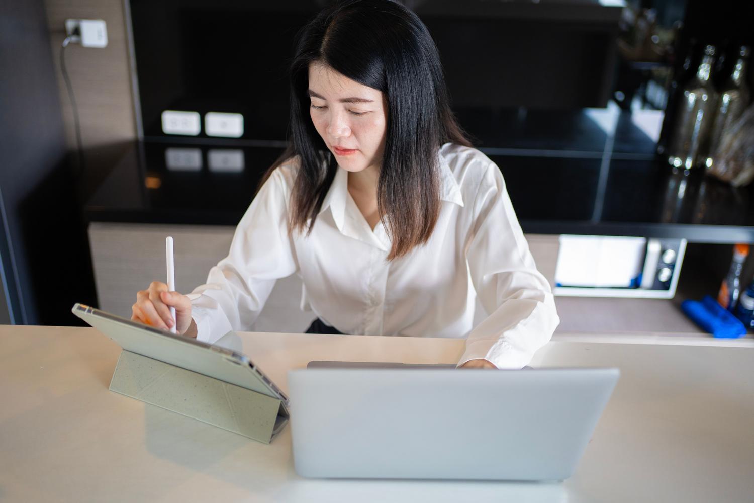 Mujer concentrada organizando información académica en una tablet y laptop en un entorno de trabajo moderno.