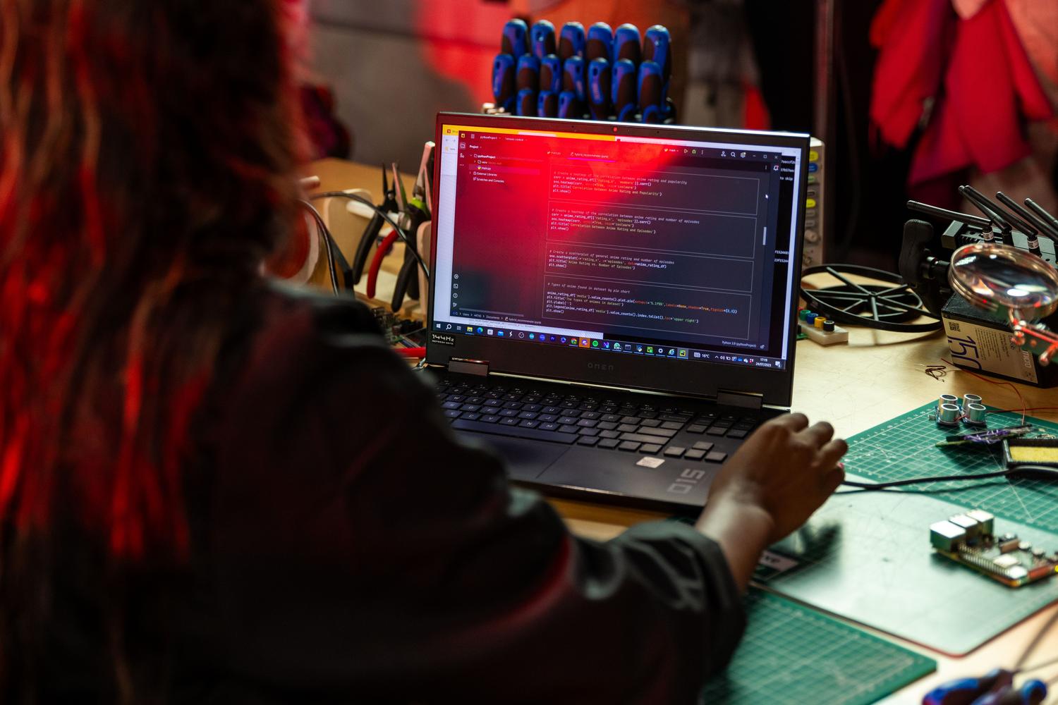 Programmerare skriver kod på en bärbar dator vid en arbetsbänk full av elektroniska verktyg.